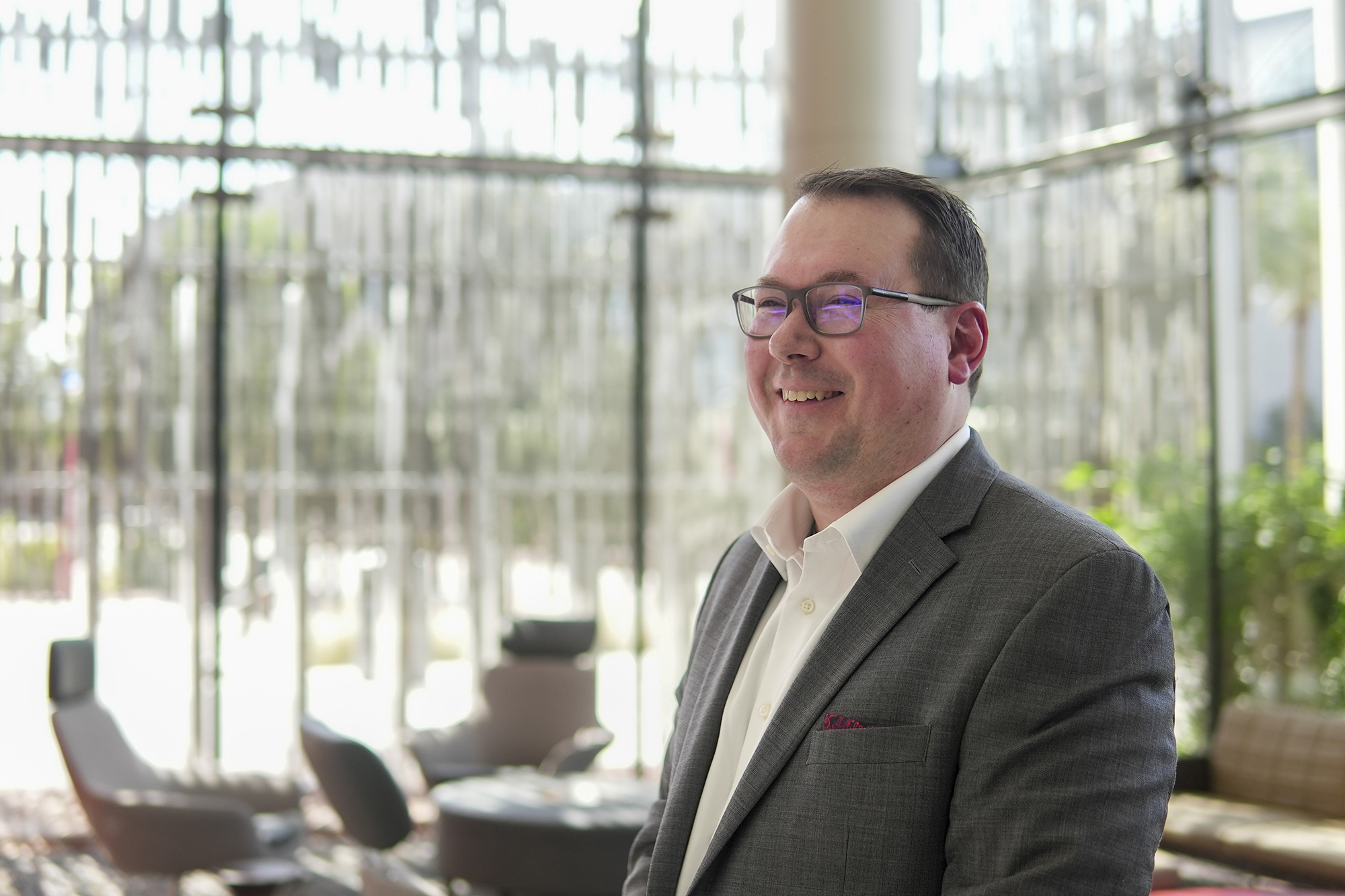 Person wearing suit jacket and glasses while inside building with large windows and lounge chairs.