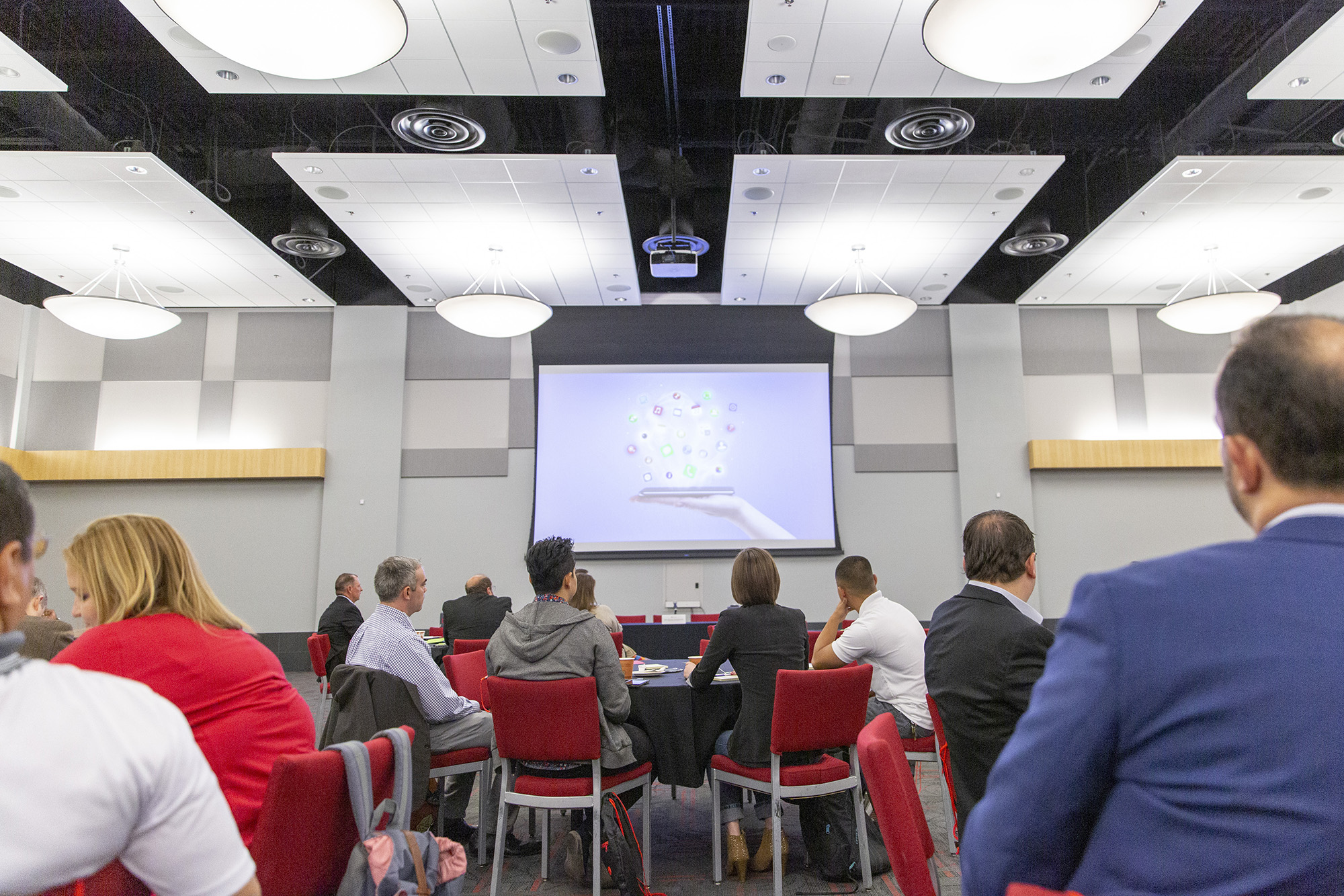 Groups of people sitting at tables looking at a projection screen on the wall.