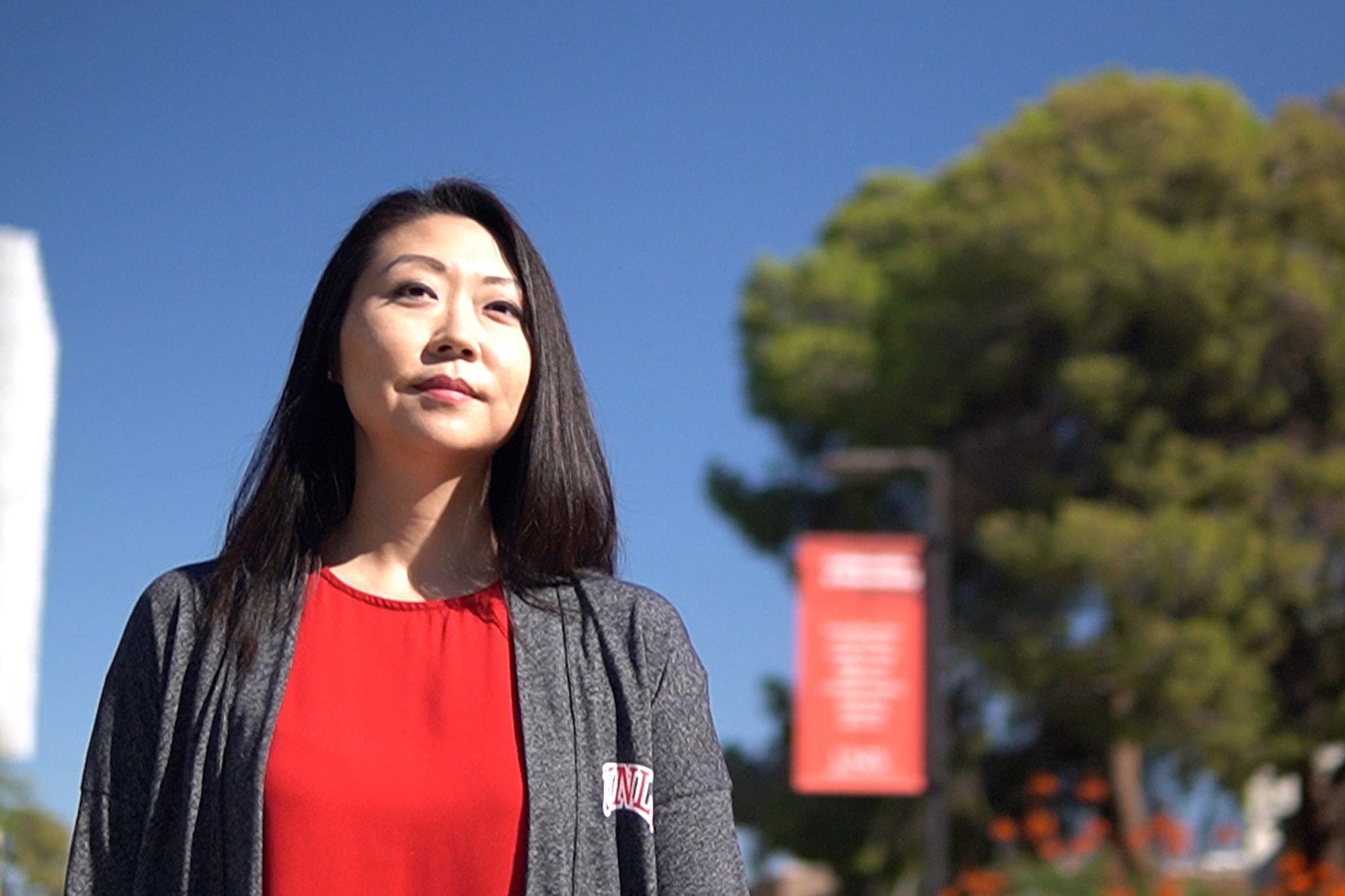 Person wearing a red shirt and gray cardigan standing outside in front of trees and red banner.