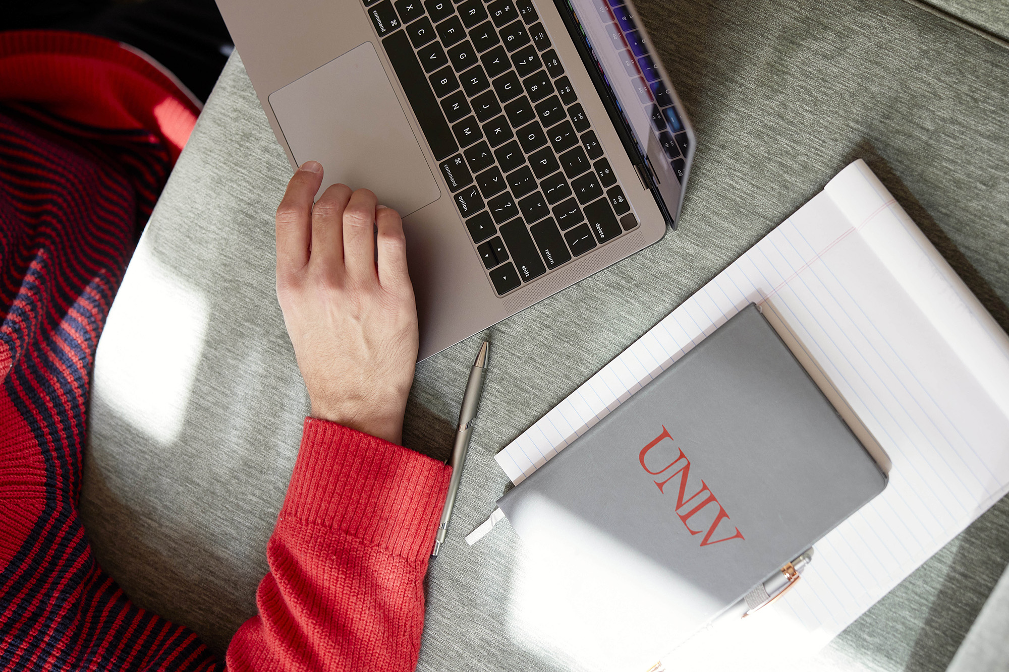 A person wearing a red and black striped sweater working on a laptop with a silver pen, a notebook with "UNLV" written on the cover placed next to their arm.