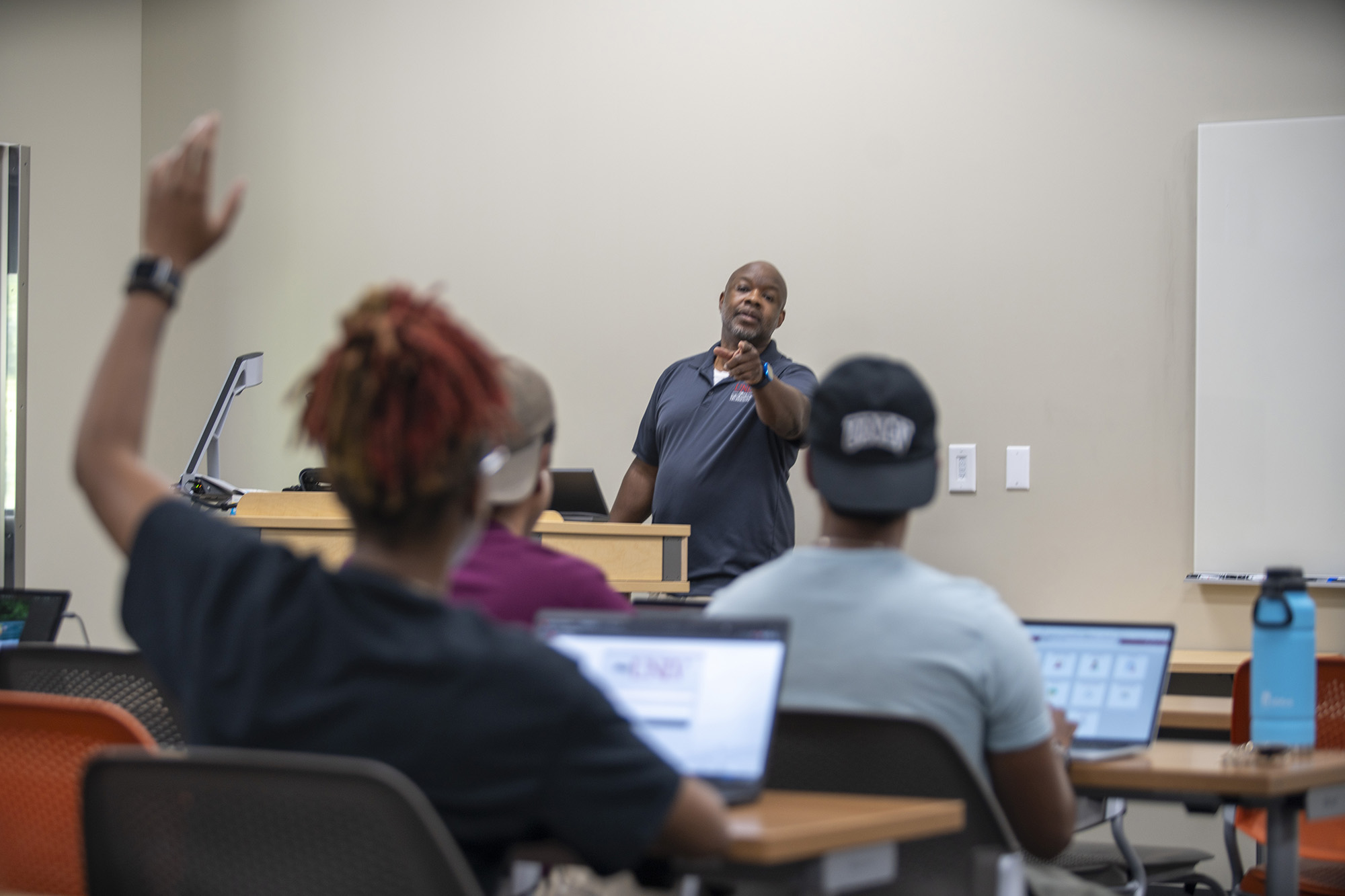 College instructor engaging with students in a modern classroom; one student has their hand raised while others work on laptops.