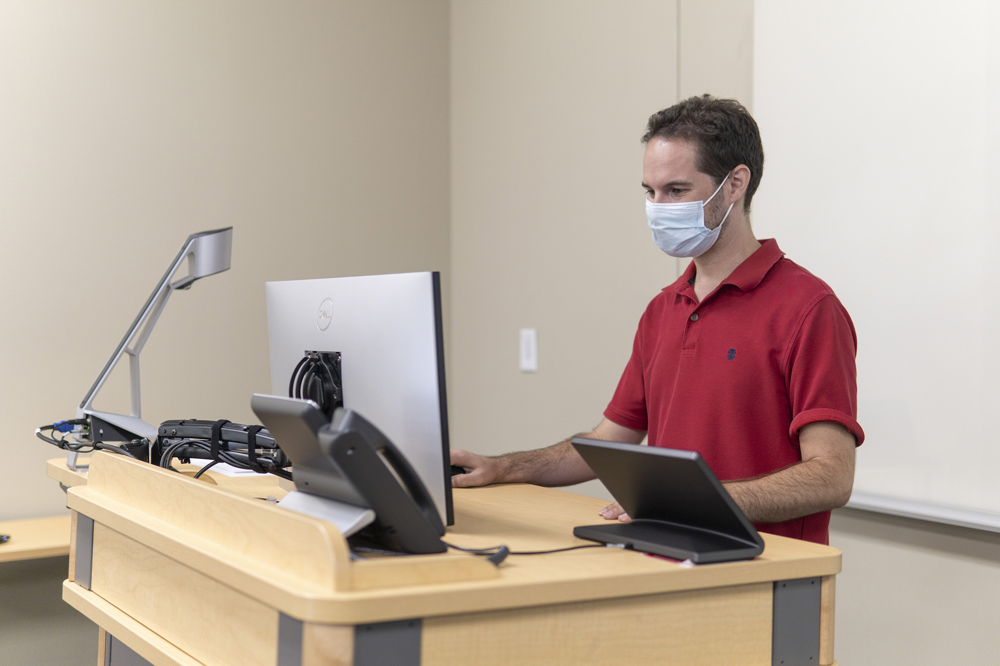 A masked staff member standing at a lectern gazes down at a computer monitor.