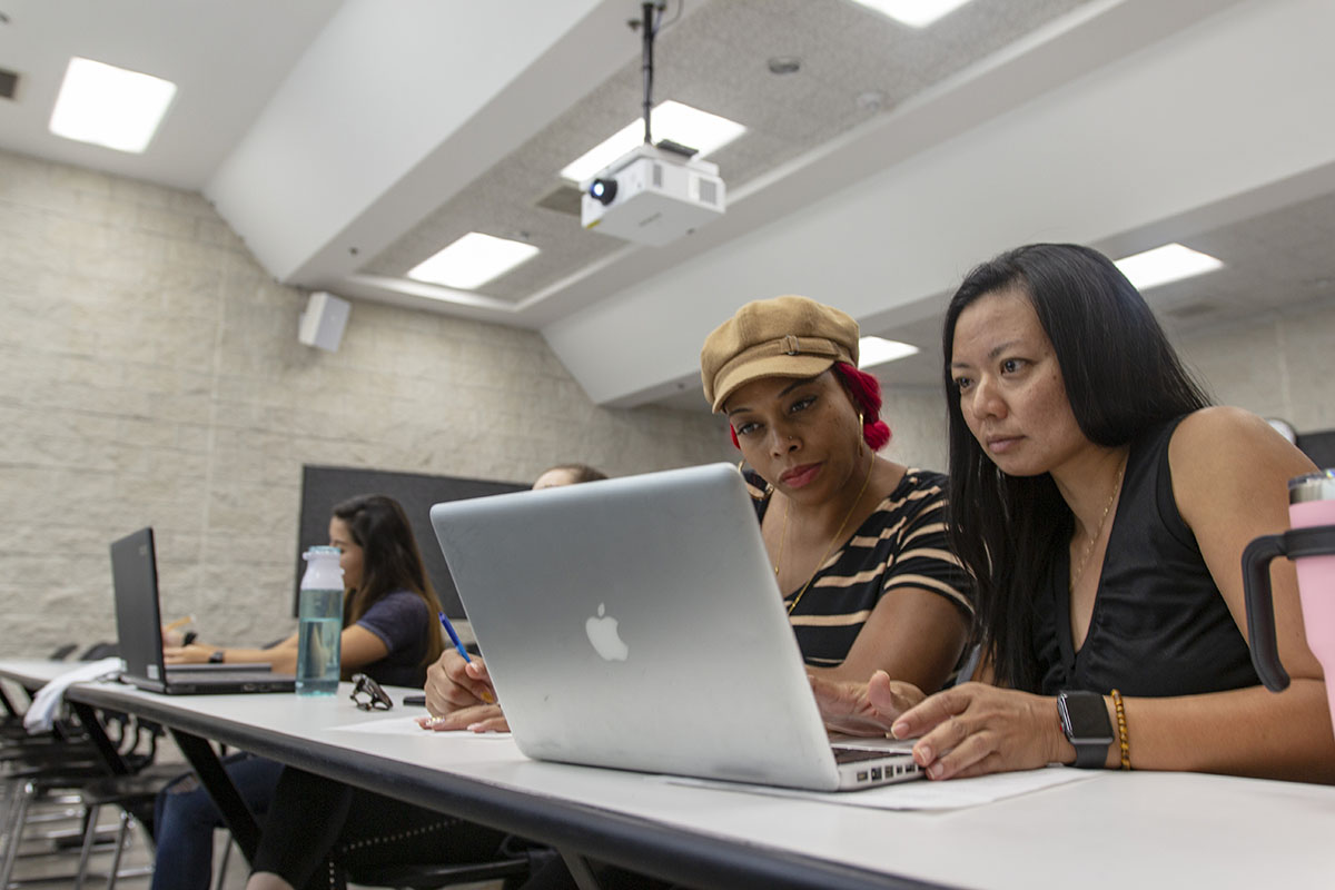 Two female staff members look down at an open Macbook.