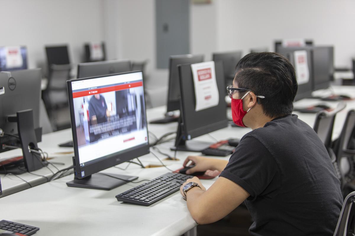A student using desktop computer in computer lab while wearing a red face covering.