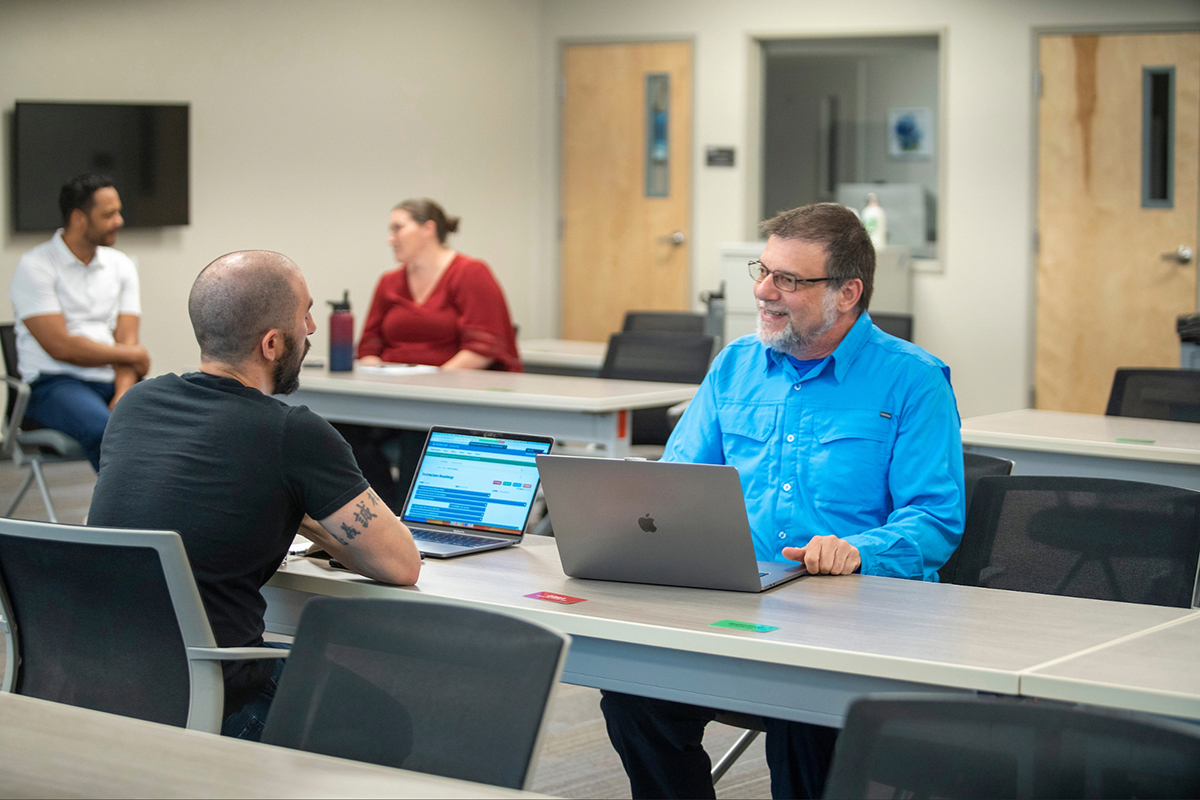 Two people talk across a table while using laptops in a classroom, with two others conversing in the background.