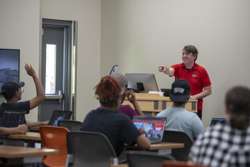 Instructor at the front of the classroom pointing at students sitting at tables 
