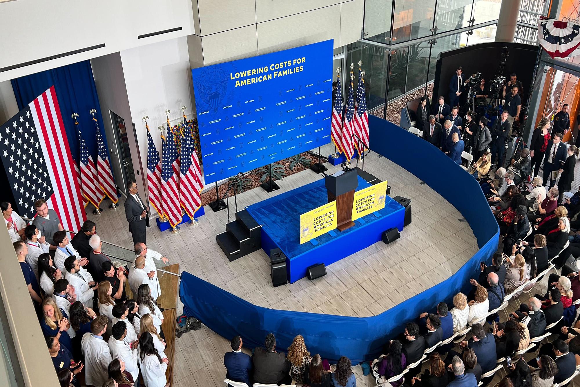A room with people standing and sitting near a podium with a banner that says lowering costs for American families