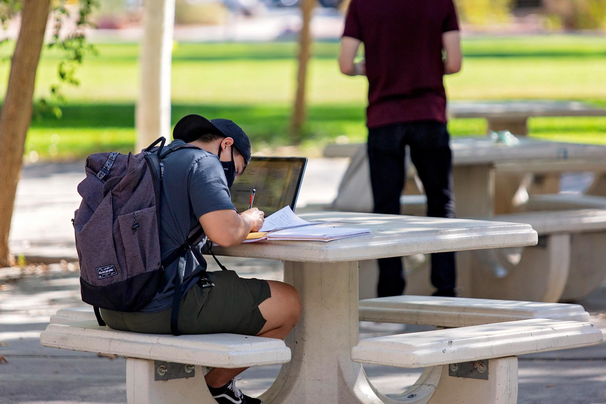 Student writing in notebook while using a laptop outside.