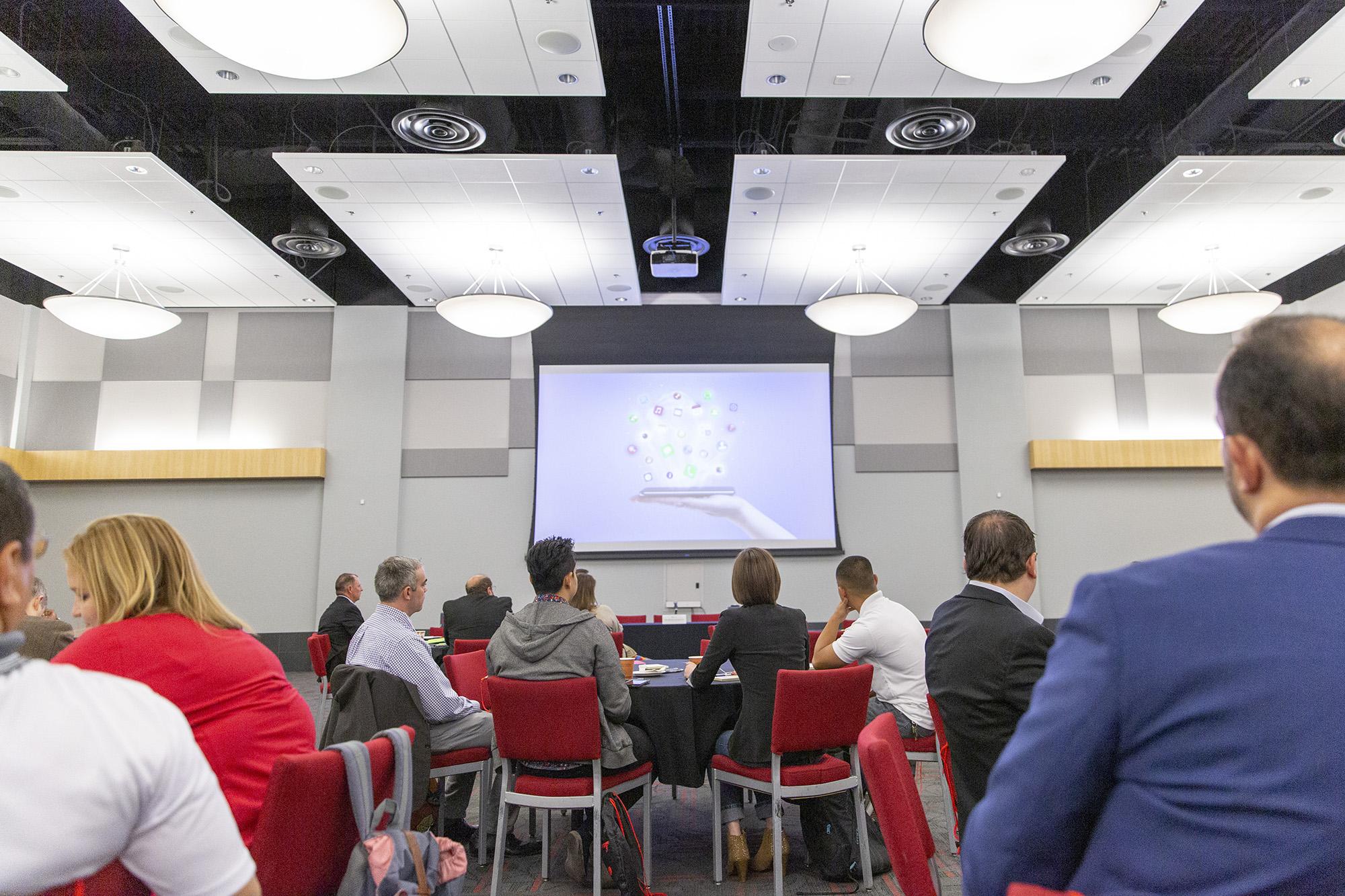 Groups of people sitting at tables looking at a projection screen on the wall.
