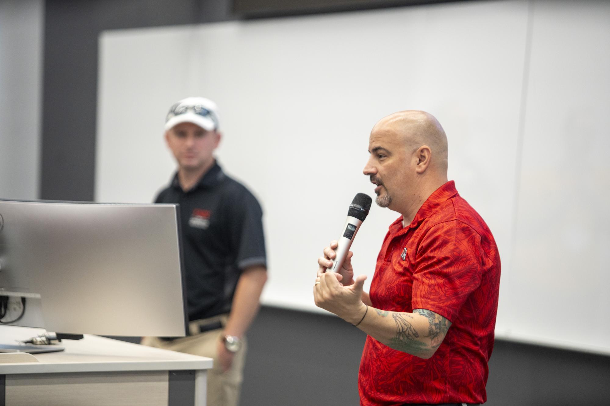 Person holding a microphone and talking with another person in the background