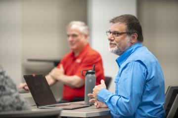 Person in blue button up shirt sitting behind laptop with person in red shirt in the background.