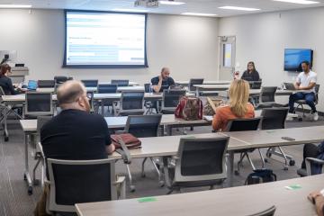 Wide shot of classroom filed with people sitting at the desks facing forward looking at a pulled down projector screen mounted in the front of the room.
