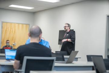 Person in black polo standing in a classroom talking to people sitting down at the desks.