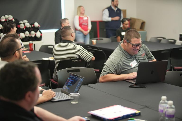 Person sitting and smiling while using a laptop computer surrounded by other people sitting at nearby tables.