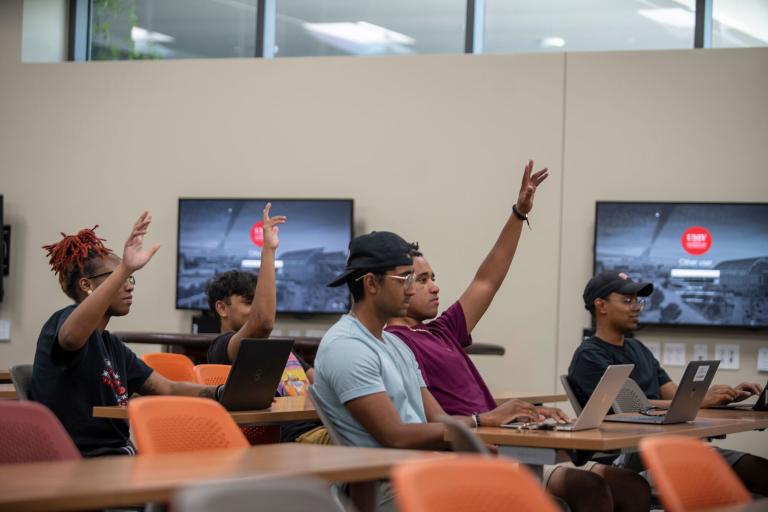A group of people sitting at tables and raising their hands in a classroom