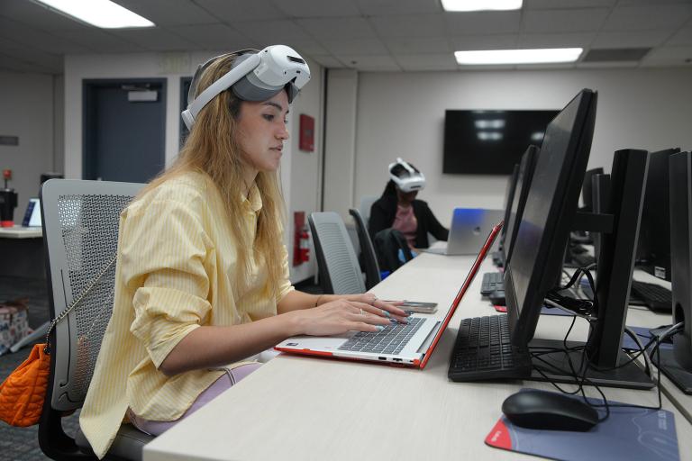 Person wearing a virtual reality headset while typing on a laptop at a desk in a computer lab, with another person using a VR headset in the background.