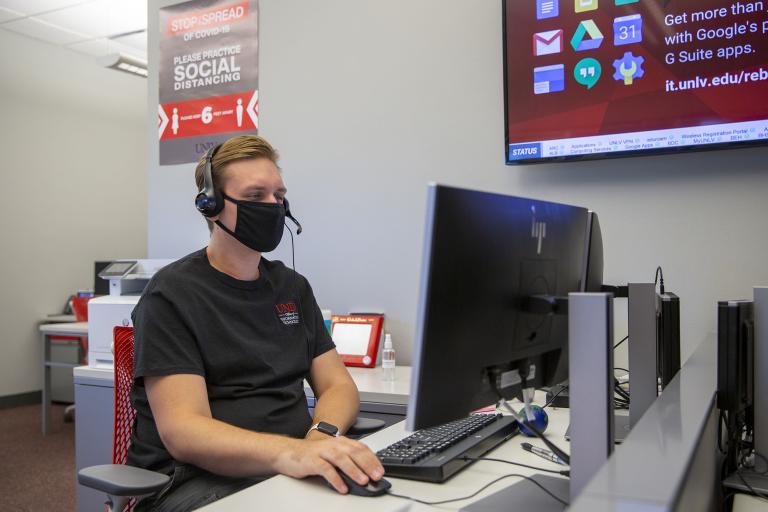 A masked IT Help Desk worker fields calls at his workstation.