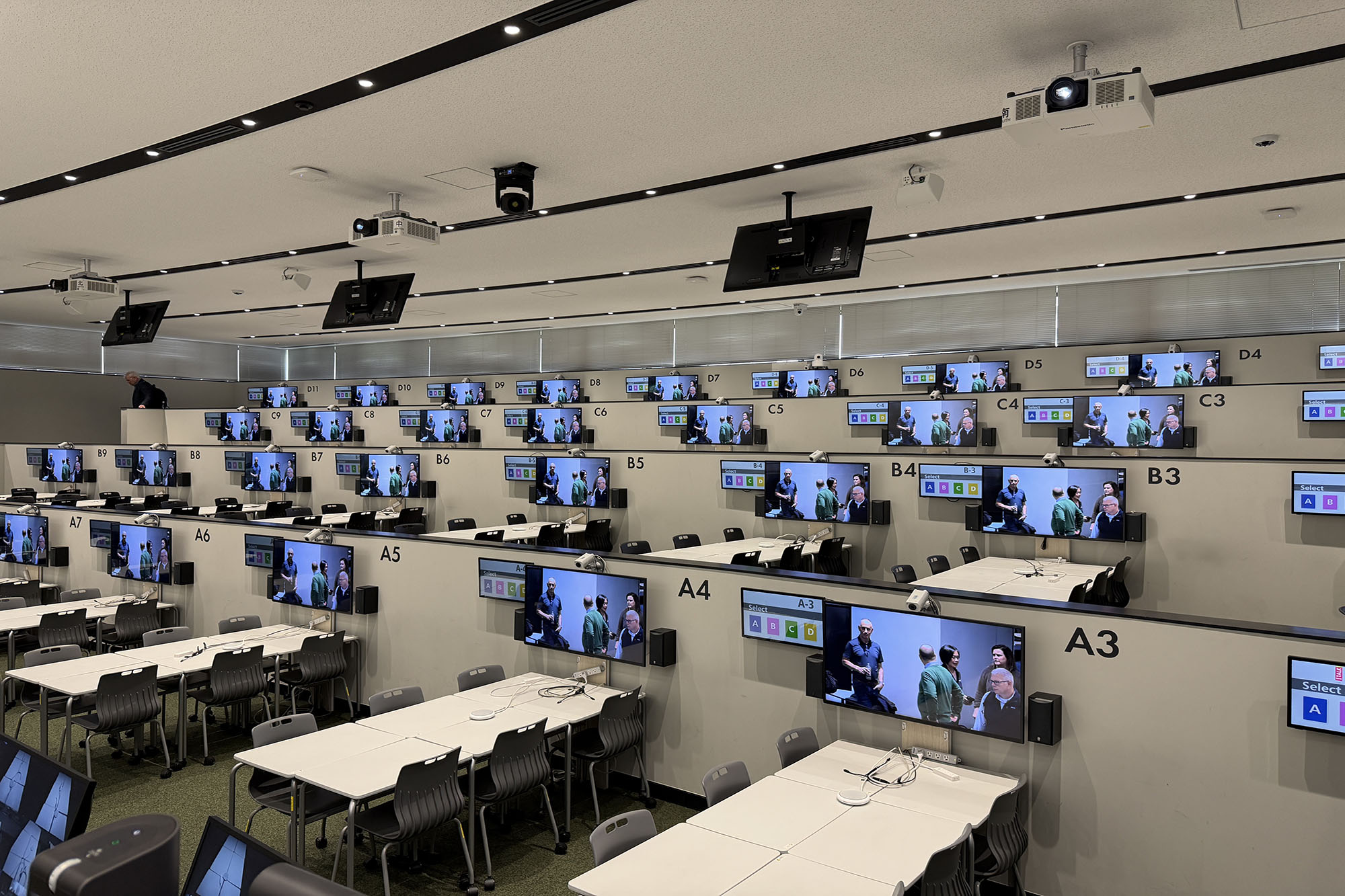 Large tiered classroom with grouped tables, ceiling-mounted projectors, and multiple wall-mounted monitors displaying instructional content.
